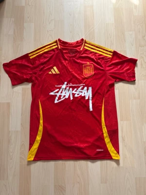 Spanien x Stüssy Hemma Kit 2025 - Stilren fotbollströja i helt nytt skick. Tröjan är inte använd en endaste sekund. Spanien tröjan kombineras med det exklusiva streetwear märket Stüssy.