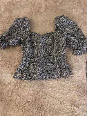 Blommig topp med puffärmar - Supersöt blommig topp med puffiga ärmar och smickrande v-ringning. Toppens småblommiga mönster går i vitt och rosa mot en mörk botten. Modellen är lätt croppad, storlek XXS-XS. Aldrig använd, köpt från HM Divided