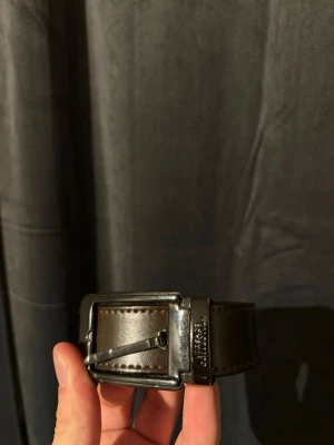 Versace Läderbälte - Stilrent brunt läderbälte från Versace. Riktigt bra skick utöver ytterst små creasningar vid ett av hålen vilket förkommer lätt vid användning. Storlek 90/105 justerbart.