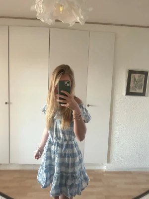 Rutig Mayoral klänning - Söt kortklänning med blå och vita rutor, offshoulder och volangdetaljer upptill. Klänningen har elastisk midja och är tillverkad i ett lätt bomullsmaterial. Perfekt för sommarens varma dagar och har en luftig passform.