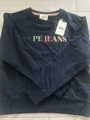 Mörkblå sweatshirt från Pepe Jeans - Mörkblå sweatshirt från Pepe Jeans med broderad logga i grönt, vitt och rosa på bröstet. Tröjan har rund halsringning och ribbade muddar vid ärmslut och nederkant. Helt oanvänd med prislappen kvar (inte provad) 💕