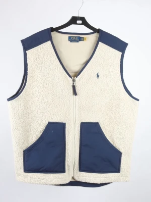 Beige/vit,  blå fleeceväst från Polo Ralph Lauren  Storlek Large - Snygg beige/vit fleeceväst från Polo Ralph Lauren med marinblå detaljer och två stora fickor framtill. Västen har dragkedja, V-ringning och den klassiska Polo-loggan broderad på bröstet. Perfekt för lager-på-lager och chill stil. Varan är begagnad och defekter kan förekomma, pris kan diskuteras, hör av dig !🤙 