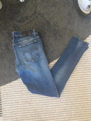 Blå slim fit jeans med stretch - Säljer ett par blå slim fit jeans med klassisk femficksdesign och snygga slitningar på framsidan. Jeansen har en mörk tvätt med ljusare partier och är tillverkade i stretchigt denim för extra komfort. Perfekt för dig som gillar en modern och smal passform.