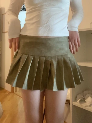 Olivgrön plisserad kortkjol - Säljer en olivgrön plisserad kortkjol med bred linning. Kjolen har en snygg och ungdomlig look med tydliga veck som ger volym och struktur. Perfekt för dig som vill ha en trendig och cool stil.