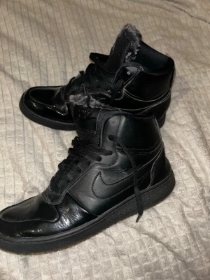 Svarta Nike sneakers med fuskpäls - Säljer ett par svarta Nike sneakers i high modell med snörning och rund tå. Skorna är i skinn och har mjuk grå fuskpäls på insidan för extra värme. Klassisk Nike-logga på sidan och svart sula. Perfekta för dig som vill ha både stil och komfort.