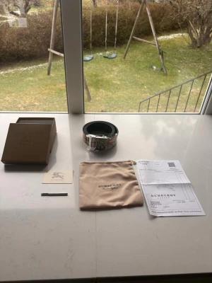 Burberry bälte med klassiskt rutmönster med kvitto - Burberry bälte i skinn med ikoniskt rutmönster i beige, brunt och rött. Snyggt silverfärgat TB-spänne framtill. Perfekt accessoar för att lyfta din outfit. Levereras med originalbox, dustbag och kvitto.