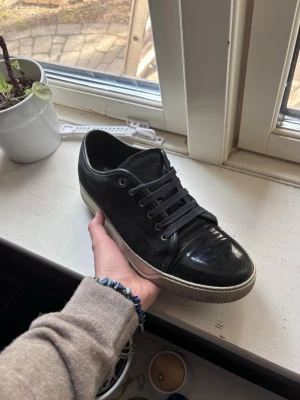 Lanvins - Säljer ett par svarta sneakers med vit sula och svarta skosnören. Skorna har en låg profil och kombinerar både mocka och blankt läder på ovandelen. Perfekt för dig som vill ha en stilren och klassisk look.
