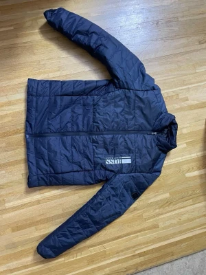 Mörkblå pufferjacka från BOSS - Snygg mörkblå pufferjacka från BOSS med quiltad design och dragkedja framtill. Jackan har ett tryckt BOSS-logo på bröstet och är tillverkad i ett lätt, isolerande material som passar perfekt för kalla vinterdagar. Stilsäker och modern look.