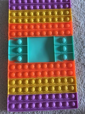 Färgglad pop it rektangel spel - Säljer en rektangulär pop it i silikon med färgglada ränder i lila, gul, orange och turkos. Perfekt för stressavlastning eller som leksak. Den har en unik design med en tom ruta i mitten och bubblor runt om. Det är ett spel. Man behöver en tärning och så slår man den och poppar så många som tärningen visar. Den som blir klar med sin  sida först vinner.