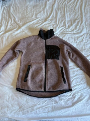 Brun fleecejacka med svarta detaljer - Säljer en brun fleecejacka med svarta detaljer och dragkedja. Jackan har hög krage, svarta fickor med dragkedja och en svart bröstficka. Perfekt för lager-på-lager och chill dagar. Materialet är mjukt och värmande.