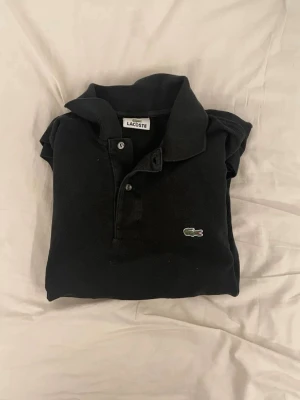 Svart polotopp från Lacoste - Klassisk svart polotopp från Lacoste med den ikoniska krokodilloggan broderad på bröstet. Toppens krage och knappar ger en stilren look som passar till många outfits. Materialet är mjukt och bekvämt, perfekt för dig som gillar enkel och tidlös stil.