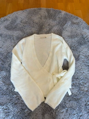 Vit omlottkofta från 157 - Vit stickad omlottkofta från 157 med knytband i sidan och djup v-ringning. Den är helt ny