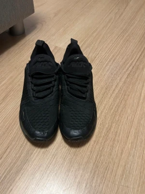 Nike Air Max 270 helsvarta sneakers - Snygga helsvarta Nike Air Max 270 sneakers med mesh-överdel och synlig Air-enhet i hälen. Skorna har en sportig siluett, svarta skosnören och diskreta loggor på plösen och sidan. Perfekta för dig som gillar stilrena och bekväma sneakers.