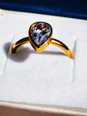 Guldfärgad ring med droppformad sten - En elegant guldfärgad ring med en stor, droppformad klar sten som gnistrar i ljuset. Ringen har en enkel och stilren design med en slät band och passar perfekt för dig som vill ha något som sticker ut. Levereras i ask.