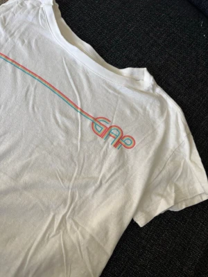 Vit GAP t-shirt med retrotryck - Snygg vit t-shirt från GAP med ett färgglatt retroinspirerat tryck i blått, rosa och gult över bröstet. Klassisk passform och korta ärmar. Perfekt till jeans eller shorts för en avslappnad look.
