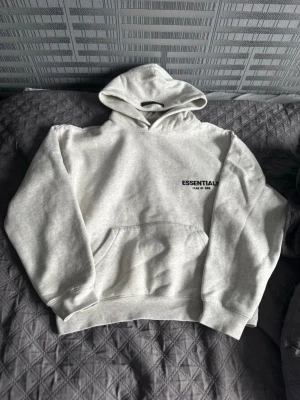 Essentials Fear of God grå hoodie - Snygg grå hoodie från Essentials Fear of God med stor svart logga på ryggen och mindre tryck på bröstet. Klassisk passform med huva och känguruficka framtill. Mjuk bomullsblandning som är perfekt för chill dagar.