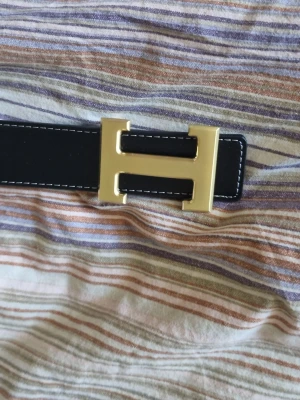 Svart skärp med H guldspänne - Snyggt svart skärp från He med ett stort H-format spänne i guld. Skärpet har vita sömmar längs kanterna och är gjort i slätt skinn. Perfekt för att lyfta vilken outfit som helst.