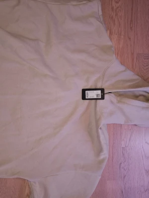 Beige hoodie med huva, storlek L - Säljer en stilren beige hoodie med huva i storlek L. Hoodien har en enkel och clean design utan tryck eller logga, perfekt för dig som gillar minimalistiskt mode. Tillverkad i mjukt material som känns skönt mot huden. Passar perfekt till jeans eller sweatpants.