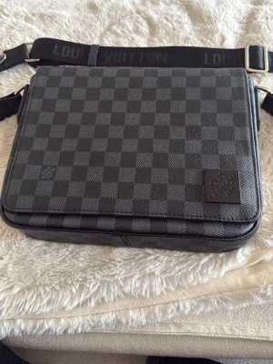 Louis Vuitton axelremsväska svart/grå - Säljer en snygg axelremsväska från Louis Vuitton i svart och grå rutigt mönster. Väskan har justerbar rem med logotyp, en yttre ficka med dragkedja och detaljer i läder. Perfekt för att bära dina essentials.