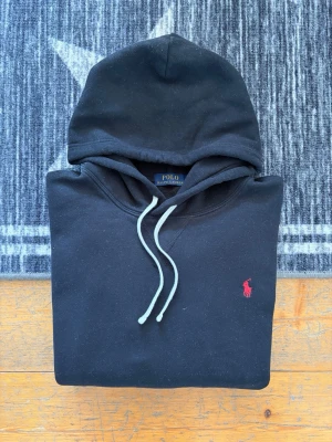 Svart Polo Ralph Lauren Hoodie - Hej! 👋 Säljer en klassisk svart Polo Ralph Lauren hoodie. Den har den ikoniska röda pony-loggan broderad på bröstet – den mest tidlösa färgkombinationen som aldrig går ur stil! 🐎 Tröjan är i mycket fint skick, djupsvart i färgen och nästintill fri från noppor. Den är 100% äkta med alla tvättlappar kvar och har QR-kod för enkel verifiering av äkthet, vilket gör det tryggt och smidigt för dig som köpare ✅ Ett premium-basplagg av högsta kvalitet. Fraktar snabbt! 🚛📦💬💯 