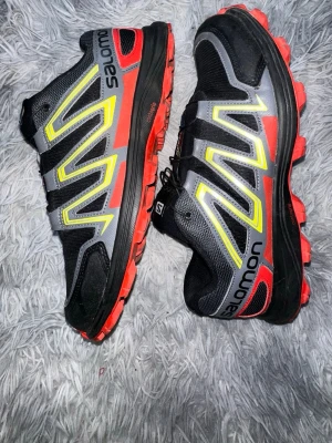 Salomon Speedtrak svarta/röda kängor - Säljer ett par Salomon Speedtrak kängor i svart och rött med gula detaljer. Skorna har grov sula för bra grepp, meshpaneler för ventilation och snabbsnörning. Perfekta för dig som gillar outdoor och vill ha något som sticker ut.
