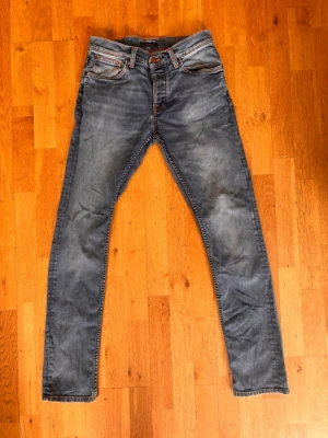 Nudie grim tim jeans - Säljer ett par blå grim tim jeans med snygga slitningar och tvättade detaljer. Jeansen har klassiska fem fickor och orangea sömmar. Perfekt för dig som gillar en avslappnad och trendig look. Storlek w31 l34.