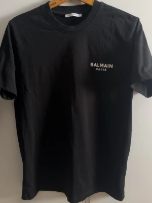 Svart Balmain Paris t-shirt - Säljer en svart t-shirt från Balmain Paris med vit logotyp på bröstet. Klassisk rund hals och korta ärmar, perfekt för dig som gillar stilrena designer. Materialet är mjukt och känns skönt mot huden. Sitter mer som S/M trots att det står xs