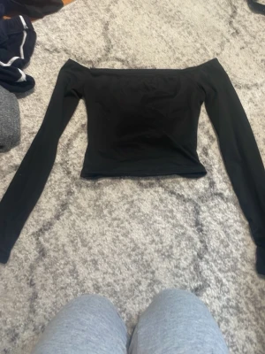 Svart offshoulder långärmad topp - Säljer en svart offshoulder topp med långa ärmar. Toppens stil är enkel och slim, perfekt för dig som gillar en clean look. Materialet känns mjukt och stretchigt, troligen polyester eller bomull. Passar dig som vill ha något stilrent och trendigt.