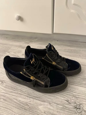 Blåa sneakers från Giuseppe Zanotti - Snygga sneakers i mörkblå sammet och svart lack från Giuseppe Zanotti. Skorna har guldfärgade dragkedjor på sidorna, svarta skosnören och en robust sula. Ikonisk logga i guld på plösen ger extra lyxig känsla.