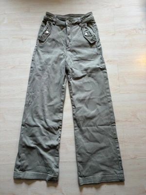 Snygga jeans - Snygga beige/gröna chinos med vida ben och coola fickor med knappar framtill. Byxorna har hög midja och klassisk design, perfekta för en avslappnad och trendig look. Materialet är mjukt och bekvämt, passar dig som gillar stilrena och tidlösa plagg.