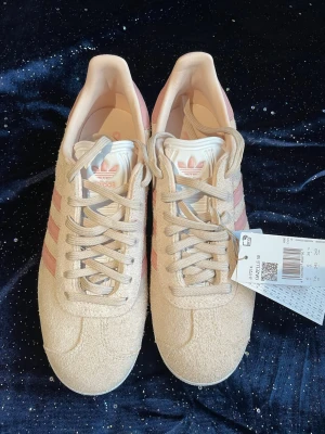 Adidas Gazelle beige/rosa sneakers - Snygga Adidas Gazelle sneakers i beige mocka med klassiska rosa ränder och vit sula. Skorna har lågt skaft, snörning och diskret Adidas-logga på plösen och sidan. Perfekta för dig som gillar retrostil och vill ha något unikt till din outfit.