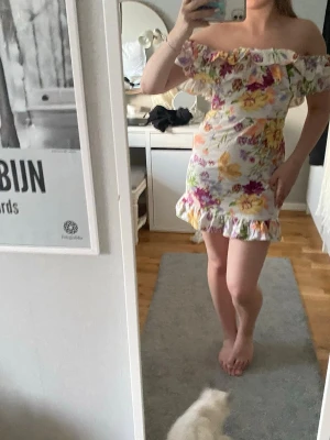 Blommig offshoulder kortklänning - Söt kortklänning med offshoulder och volangdetaljer. Klänningen har ett färgglatt blommigt mönster i rosa, gul, lila och grönt på vit botten. Perfekt för sommarens alla tillfällen, med en feminin och ungdomlig look.