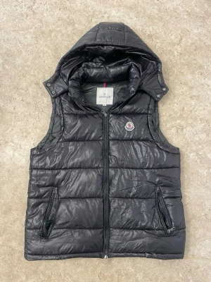 Svart quiltad dunväst från Moncler - Snygg svart dunväst från Moncler med quiltad design och avtagbar huva. Västen har två dragkedjeförsedda fickor framtill och klassisk Moncler-logga på bröstet. Perfekt för lager-på-lager och håller dig varm under kyliga dagar.