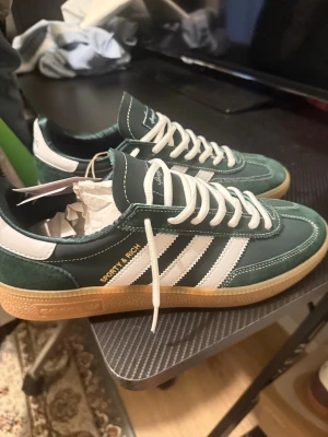 Sporty & Rich x Adidas gröna sneakers - Mörkgröna sneakers från Adidas i samarbete med Sporty & Rich. Skorna har klassiska vita ränder på sidorna, vit häl och vita skosnören. Ovandelen är i mocka och läder, med gummisula i beige. Snygg retrostil med gulddetaljer på sidan.