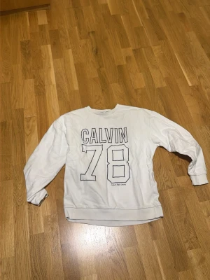 Vit sweatshirt Calvin Klein Jeans - Vit sweatshirt från Calvin Klein Jeans med trycket 'CALVIN 78' i svart på framsidan. Tröjan har rund halsringning, långa ärmar och ribbade muddar vid ärmslut och nederkant. Perfekt för en avslappnad och sportig stil.