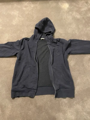 Mörkblå zip-hoodie med broderad detalj - Mörkblå hoodie med dragkedja och huva. Broderad siffra på bröstet och två stora fickor framtill. Mjuk bomullskänsla och ribbade muddar vid ärmslut och nederkant. Perfekt för chill dagar och enkel att matcha med jeans eller joggers.