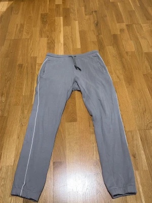 Grå mjukisbyxor med vita detaljer - Säljer ett par grå mjukisbyxor med vita piping längs sidorna. Byxorna har dragsko i midjan och två sidofickor. Perfekta för chill eller träning. Materialet är mjukt och bekvämt, och passformen är avslappnad med resår vid benslut.