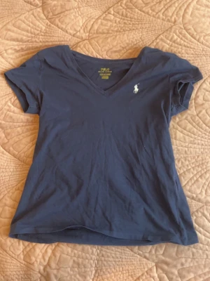 Blå v-ringad topp från Ralph Lauren - Snygg blå topp från Polo Ralph Lauren med v-ringning och klassisk logga broderad på bröstet. Tillverkad i mjuk bomull och har korta ärmar. Perfekt för en avslappnad och stilren look.