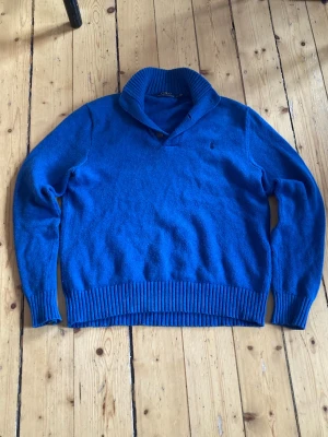 Blå stickad tröja från polo Ralph lauren med sjalkrage - Säljer en blå stickad tröja från Polo Ralph Lauren med sjalkrage och knappdetalj vid halsen. Tröjan har ribbstickade muddar och nederkant, samt diskret logga på bröstet. Perfekt för dig som gillar klassisk och stilren design.