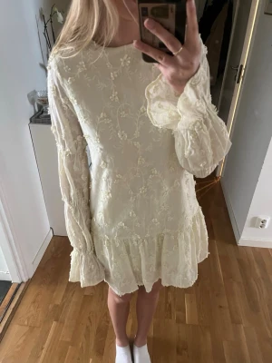 Ljusgul klänning - Ljusgul kortklänning med blommigt broderi, volangkant nedtill och långa ärmar med volangdetaljer. Klänningen har öppen rygg med knyt i nacken och är tillverkad i ett tunt, lätt material som ger en luftig känsla.