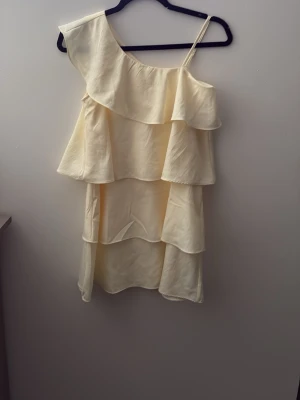 Ljus gul klänning med volanger från Gina Tricot  - Säljer en ljus gul klänning med omlott-design och flera lager volanger. Klänningen har en oneshoulder-stil med ett smalt axelband och är perfekt för sommarens alla tillfällen. Materialet känns lätt och luftigt.💓💓