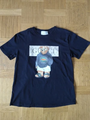 Svart t-shirt med björntryck. - Cool svart t-shirt med en stor björn i solglasögon och hoodie på framsidan. T-shirten har klassisk passform och är tillverkad i mjuk bomull. Perfekt för dig som vill sticka ut med ett exklusivt plagg.