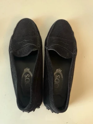 Svarta loafers från Tod's i mocka - Säljer ett par klassiska svarta loafers från Tod's i mjuk mocka. Skorna har en stilren design med pennyloafers-detalj och gummidetaljer på hälen för extra grepp. Perfekta för dig som gillar tidlös stil och komfort. Storlek 36 och oanvänd. Marinblå/svart