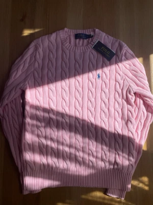 Rosa kabelstickad tröja från Polo Ralph Lauren - Säljer en ljusrosa kabelstickad tröja från Polo Ralph Lauren med rund hals och ribbade muddar. Tröjan har det klassiska broderade logotypen i blått på bröstet och är gjord i mjukt material. Perfekt för dig som gillar stilrena och trendiga plagg.