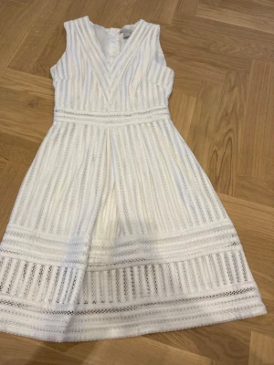 Vit ärmlös klänning med spetsdetaljer - Säljer en superfin vit klänning med v-ringning och spetsdetaljer. Klänningen är ärmlös, har ett randigt spetsmönster och en lätt utställd kjol. Perfekt för sommarens alla tillfällen! Dragkedja i ryggen för enkel påklädning.