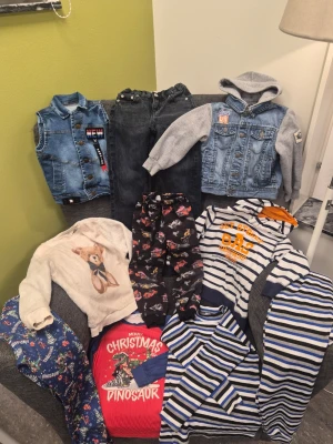 Klädpaket med blandade barnkläder 4-6 år - Parti med barnkläder. Innehåller en jeansväst, en jeansjacka med gråa ärmar och huva, ett par mörka jeans, en vit tröja med en nallebjörn, ett par svarta byxor med bilmotiv, en röd tröja med dinosauriemotiv, en vit och blårandig huvtröja samt två vit- och blårandiga långärmade tröjor. Även ett par blåa pyjamasbyxor med dinosauriemotiv.
