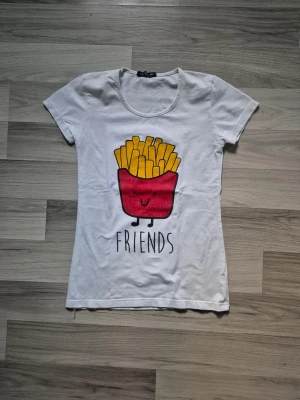 Vit t-shirt med pommes-tryck - Vit t-shirt med ett färgglatt tryck av en tecknad pommes frites och texten 'FRIENDS' på framsidan. T-shirten har rund halsringning och korta ärmar. Perfekt för dig som gillar roliga och lekfulla motiv.