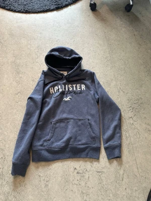 Blå Hollister hoodie med tryck - Säljer en blå hoodie från Hollister med vit text 'Hollister California' och logotyp på bröstet. Hoodien har en stor huva, ribbade muddar och en klassisk magficka. Perfekt för dig som gillar avslappnad och cool stil.