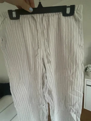 Beigevita randiga linnebyxor - Snygga linnebyxor i beigevitt med tunna vertikala ränder. Byxorna har avslappnad passform och resår i midjan för extra komfort. Perfekta för en chill och stilren look. Materialet är luftigt linne som passar bra till varmare dagar.❣️☀️😺
