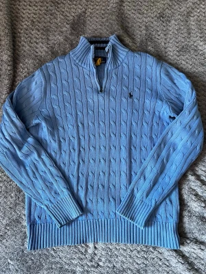 Ljusblå kabelstickad tröja Polo Ralph Lauren - Ljusblå kabelstickad tröja från Polo Ralph Lauren med halv dragkedja och klassisk broderad logga på bröstet. Tröjan har långa ärmar, ribbade muddar och är tillverkad i mjukt material. Perfekt för en stilren och avslappnad look.